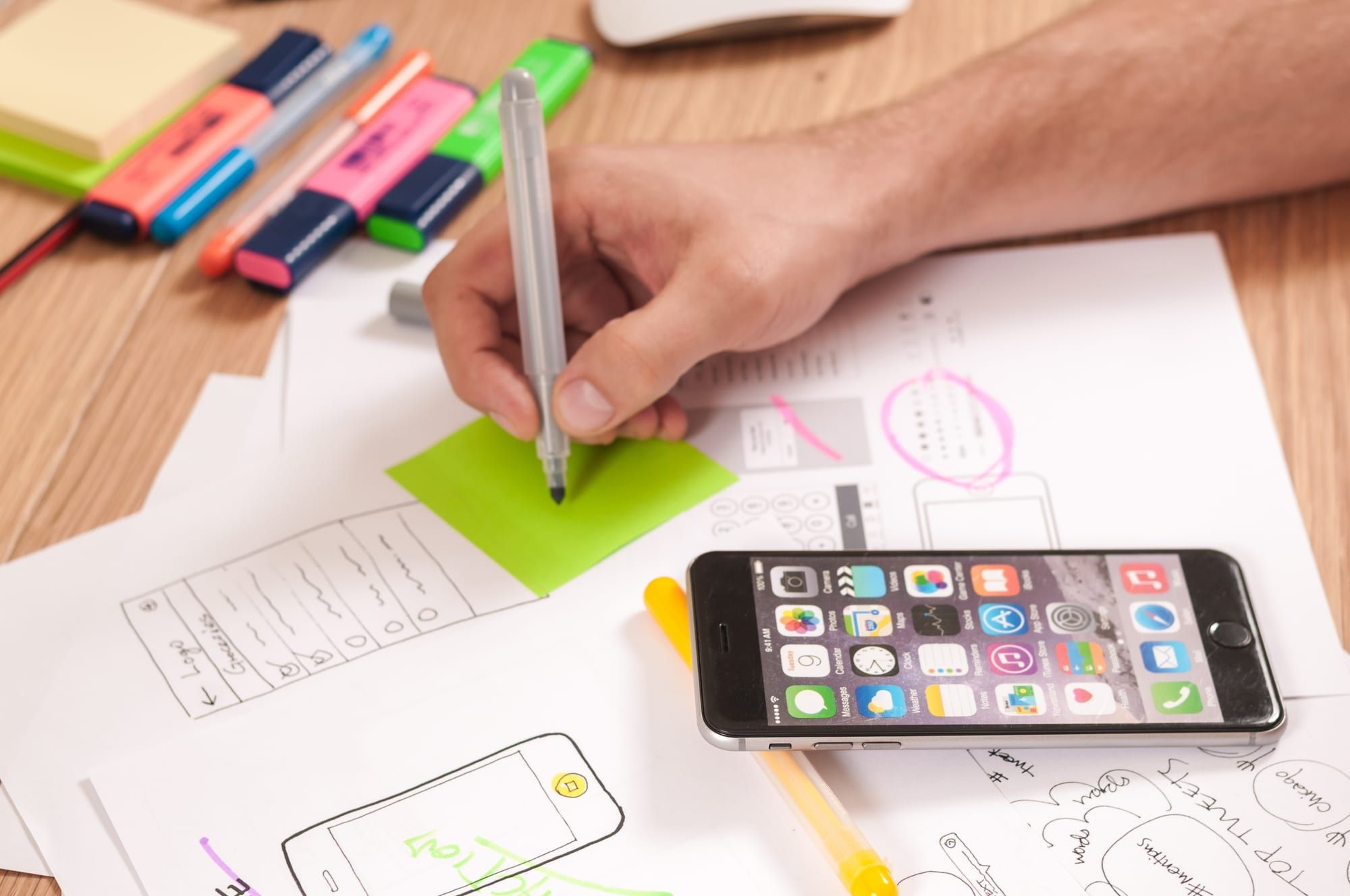 A designer sketching app wireframes on paper next to a phone and markers