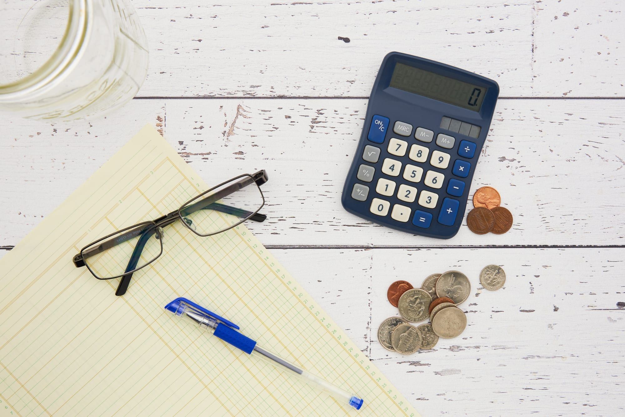 A calculator, coins, glasses and a ledger pad on a wooden table
